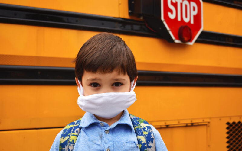 Child with mask on in front of school bus