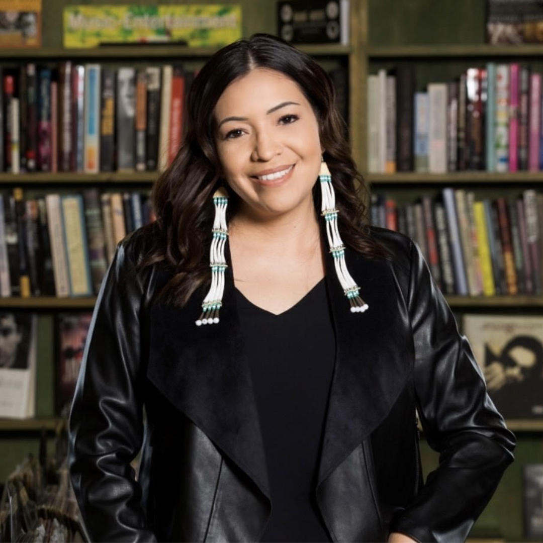 Serene Thin Elk stands in front of bookshelves, smiling, wearing black and long dentalium earrings.