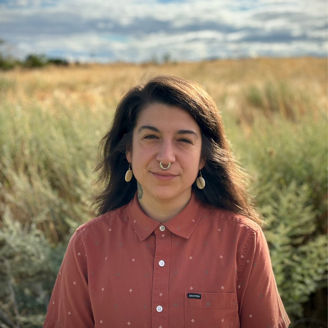 River Freemont (NABS Digital Archives Specialist) stands in front of a grassy plain wearing a red-clay colored button-up, earrings, and a septum piercing.