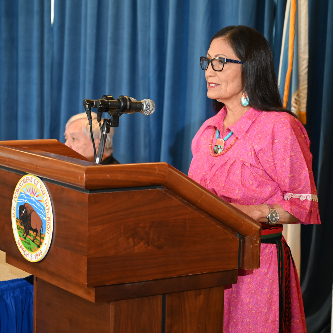 Secretary of the Interior Deb Haaland speaks at a DOI podium at the release of Vol I of the Federal Indian boarding school investigative report; Haaland wears pink and earrings.